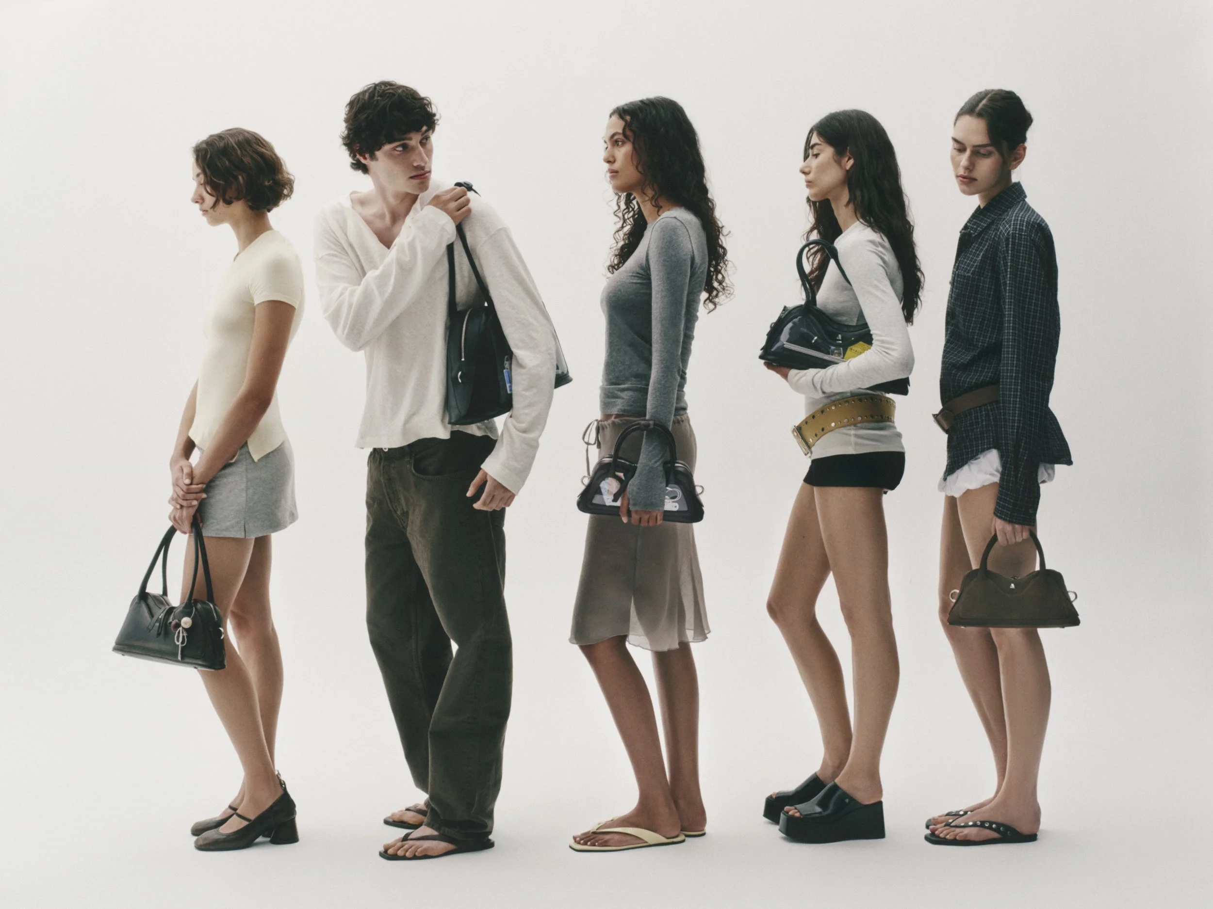 Six women standing in a row against a plain white background, facing forward with heads turned in profile, wearing casual clothing and holding bags.