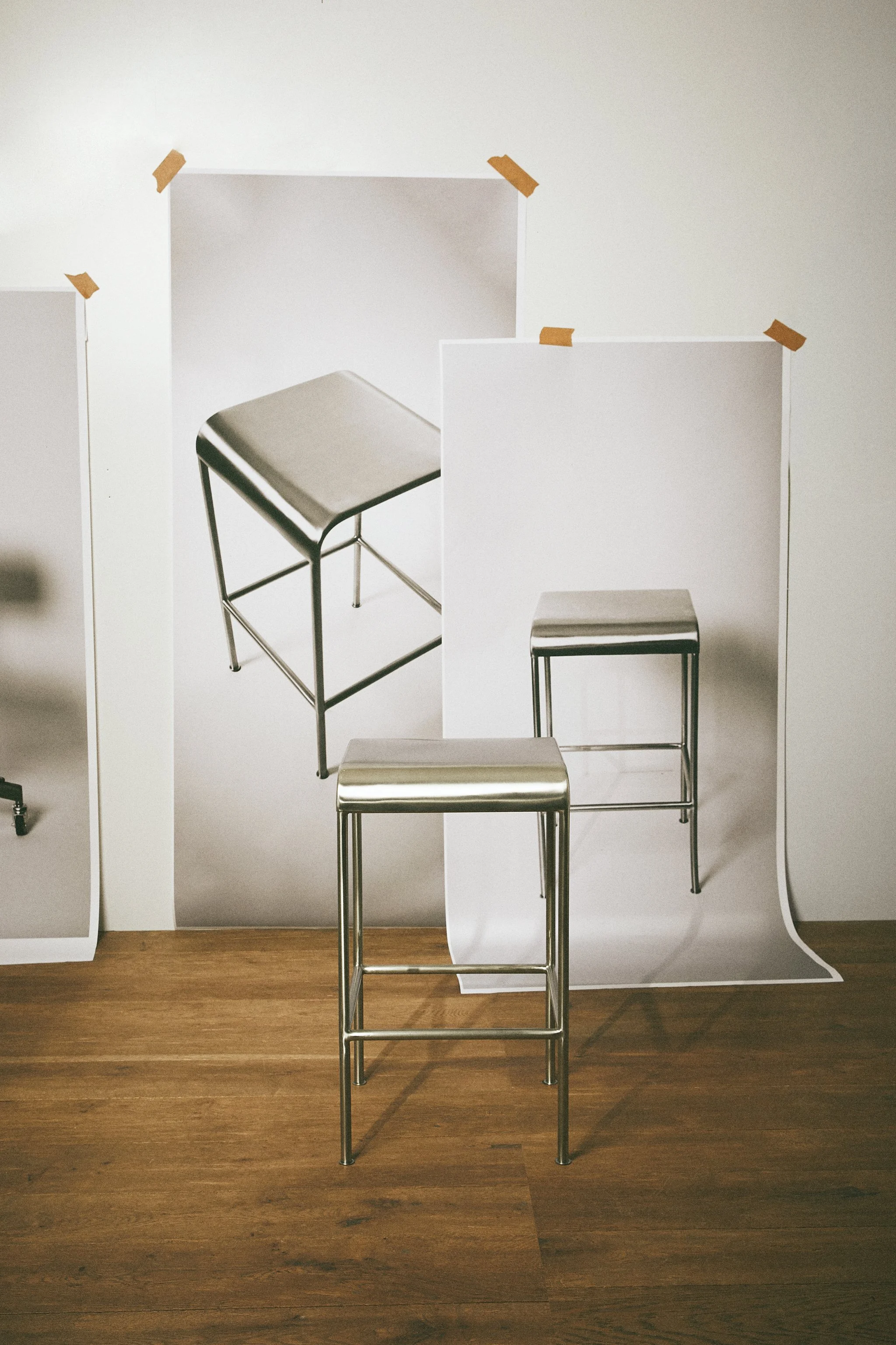 Four metal barstools with gray seats arranged in front of a white wall with cutouts and brown tape.