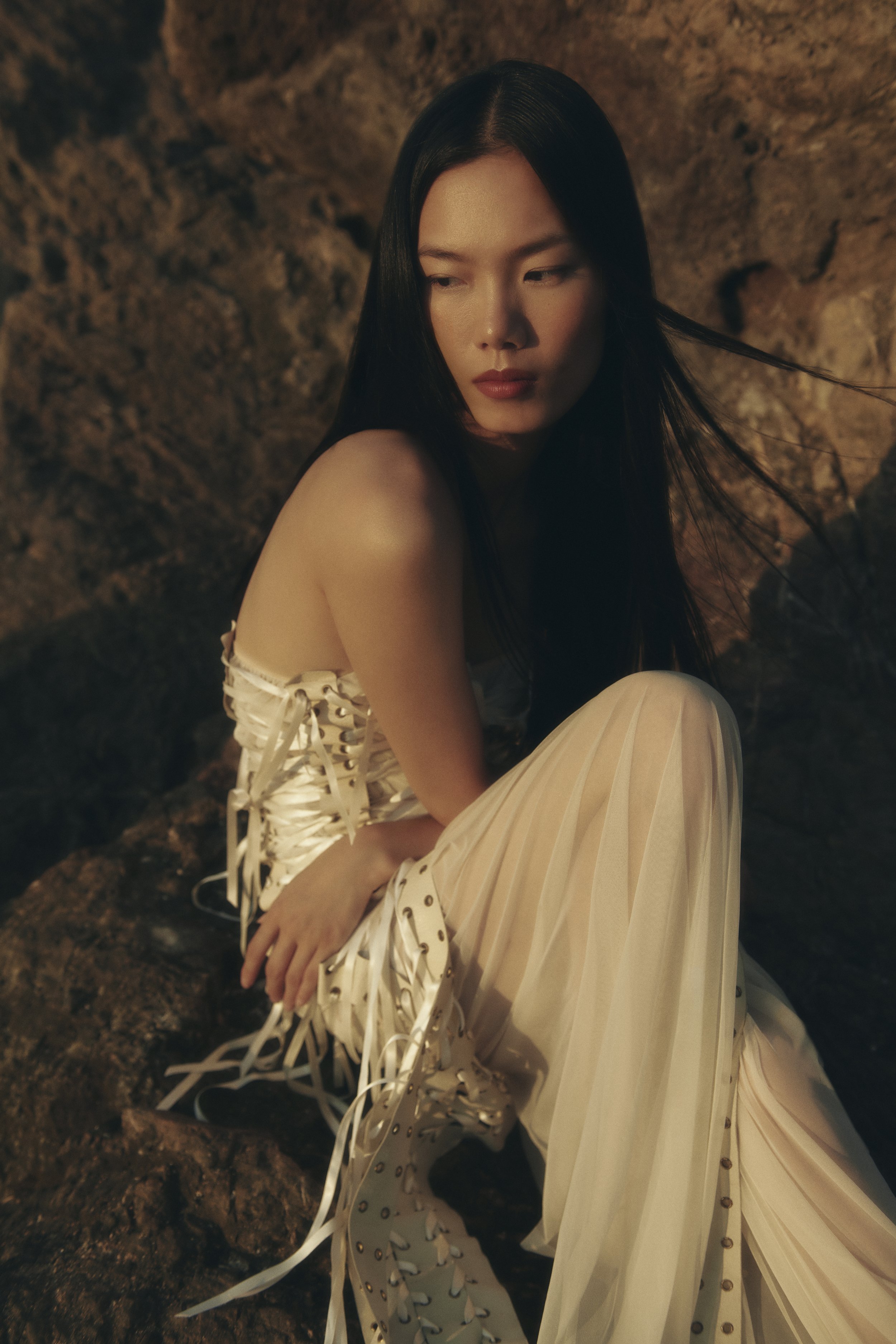 A woman with long black hair and fair skin sitting against a rocky background, wearing a cream-colored dress with lace-up details.