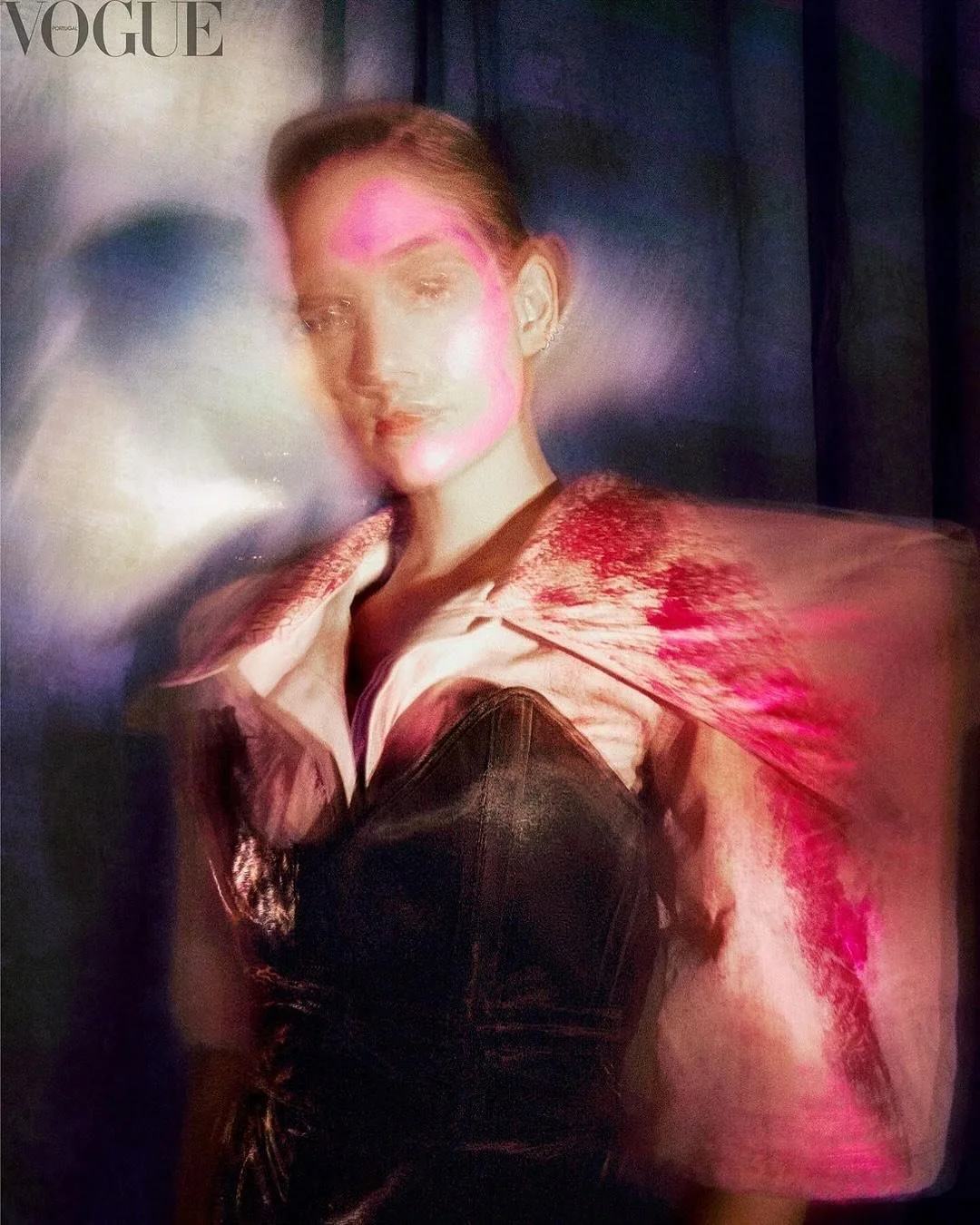 A blurred, abstract portrait of a woman with light skin, short hair, and earrings, wearing a collared shirt. The photo has bright pink and red light effects and a distorted appearance, with the Vogue logo in the top left corner.