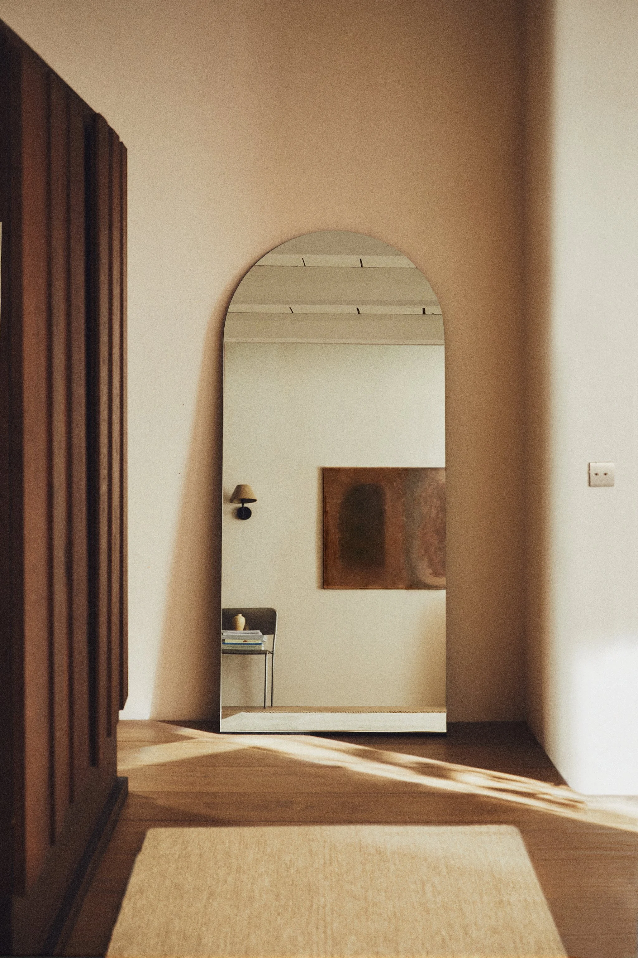 A hallway view through an arched doorway, with a mirror reflecting a small table, a lamp, and stacked books, featuring warm lighting and wooden flooring.