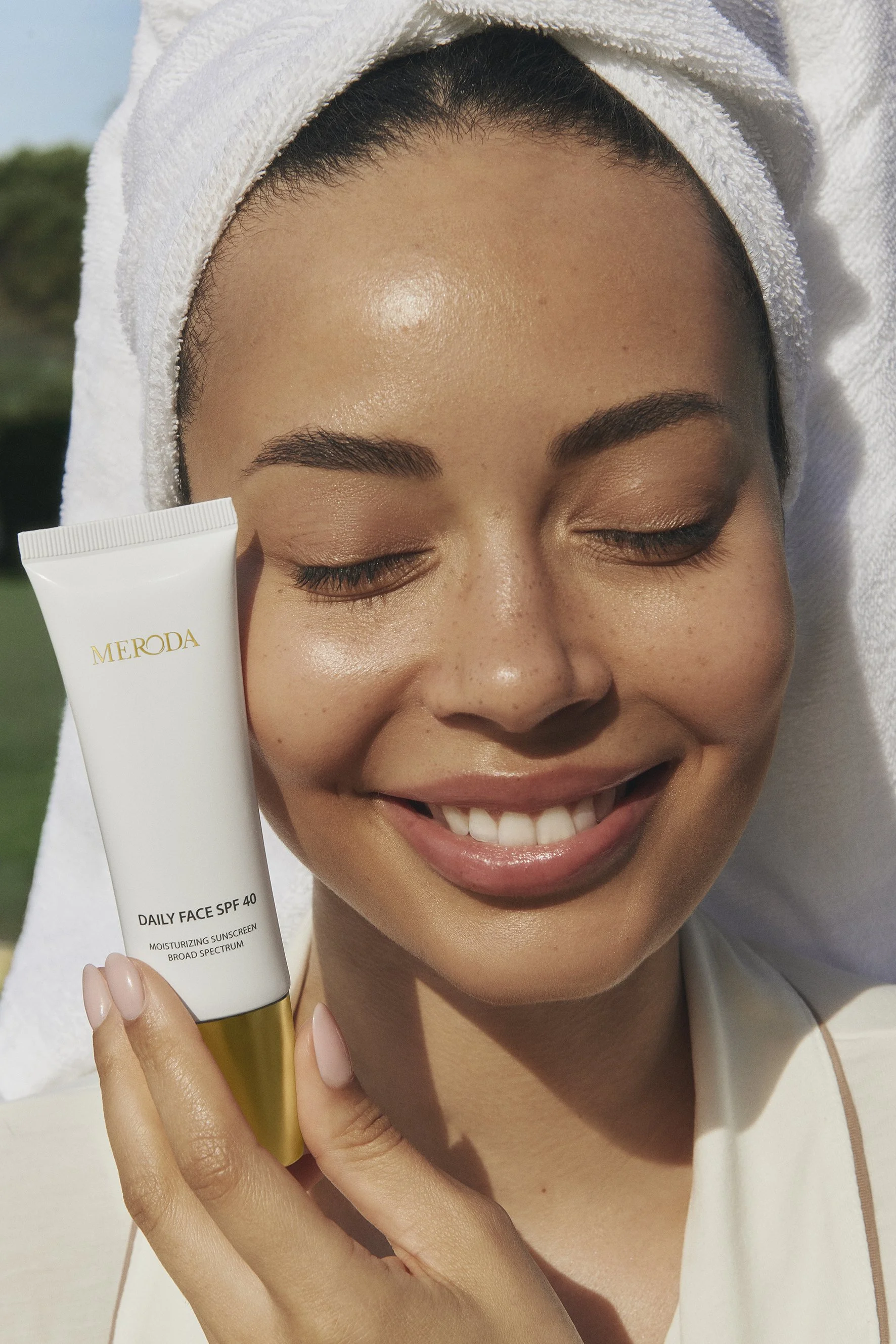 A smiling woman with closed eyes and a white towel wrapped around her head holding a tube of Meroda Daily Face SPF 40 sunscreen outdoors on a sunny day.