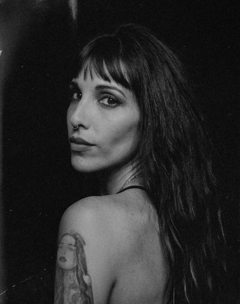Black and white portrait of a young woman with long hair and a septum piercing, looking over her shoulder at the camera. She has a tattoo on her upper arm.