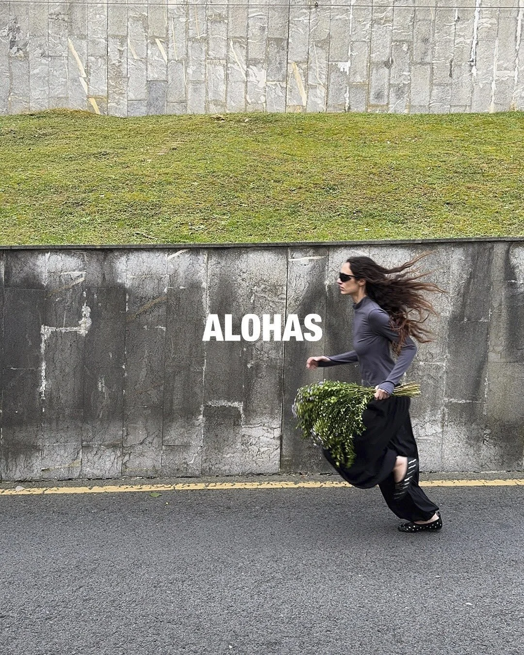 A woman with long hair wearing sunglasses, a gray top, and black pants, walking along a street while carrying a large bouquet of white flowers. The background features a stone wall and a grassy area. The word "ALOHA" is written across the middle of t