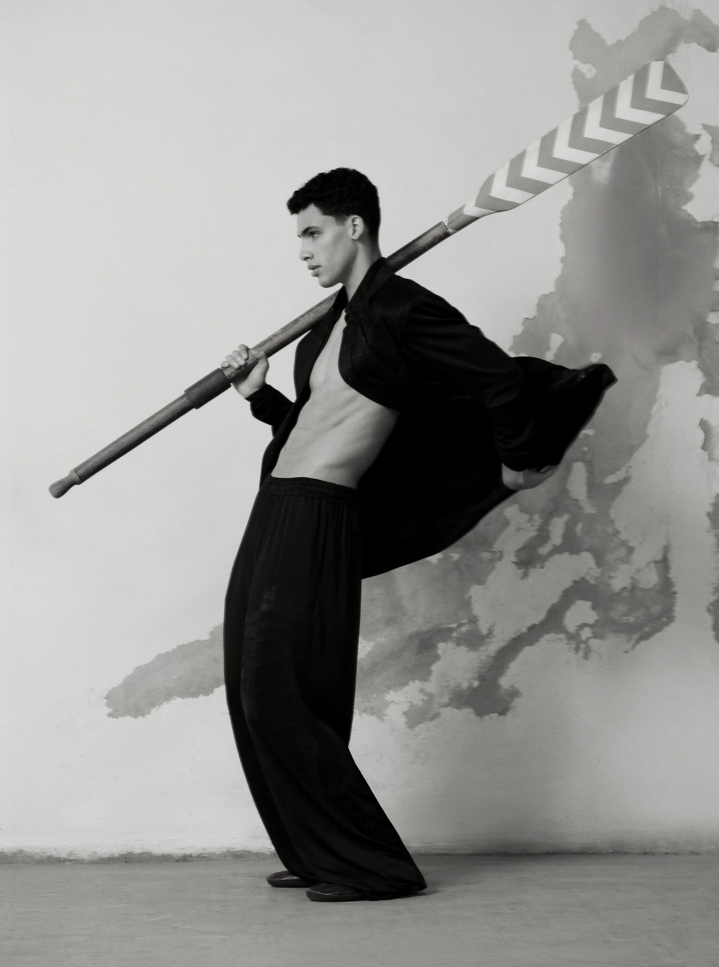A woman dressed in black pants and a light-colored shirt, with a dark jacket, is standing sideways with her back arched and head tilted forward. She is holding a long broom handle across her shoulders. Behind her is a wall with some peeling paint and