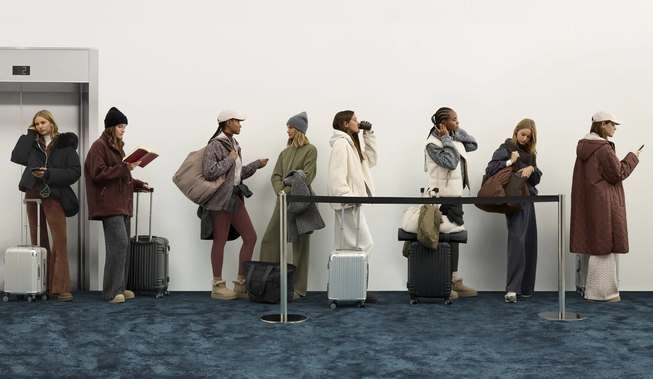 Line of women at airport check-in counter with luggage, some using phones or headphones, some talking, wearing casual winter clothing and hats.