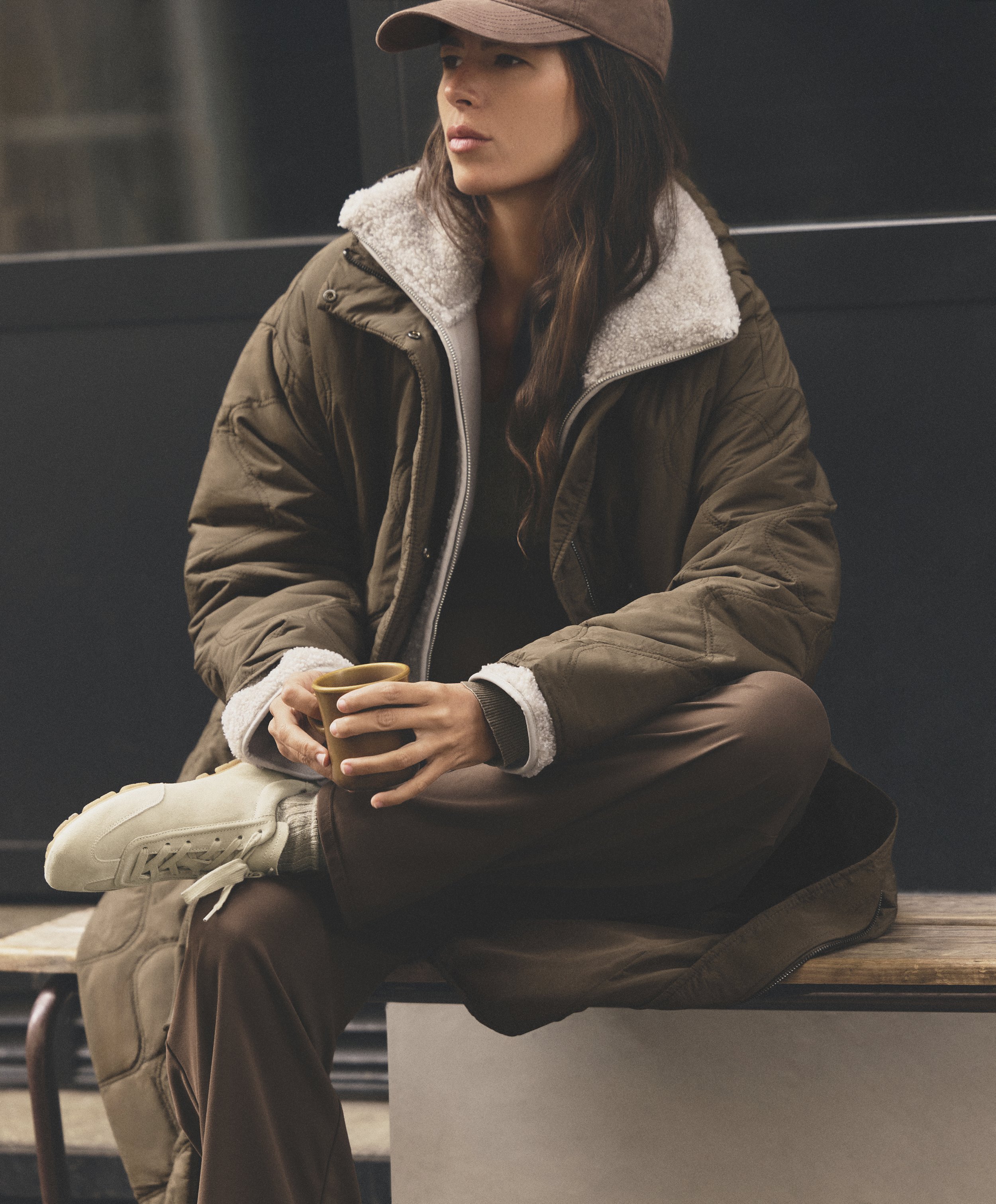 A woman in a brown jacket, beige pants, white sneakers, and a brown baseball cap sitting on a wooden bench, holding a brown mug, looking to her right.