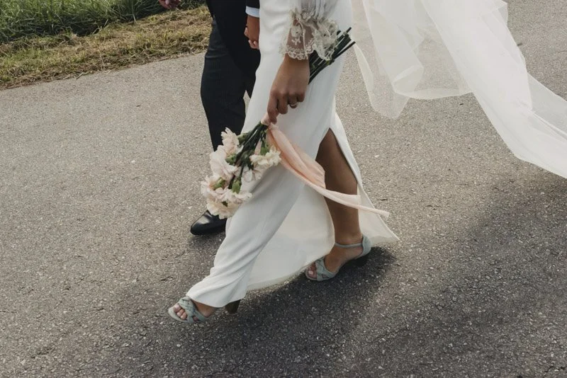 Close-up of a bride and groom holding hands, with the bride holding a bouquet of flowers, walking on a paved pathway.