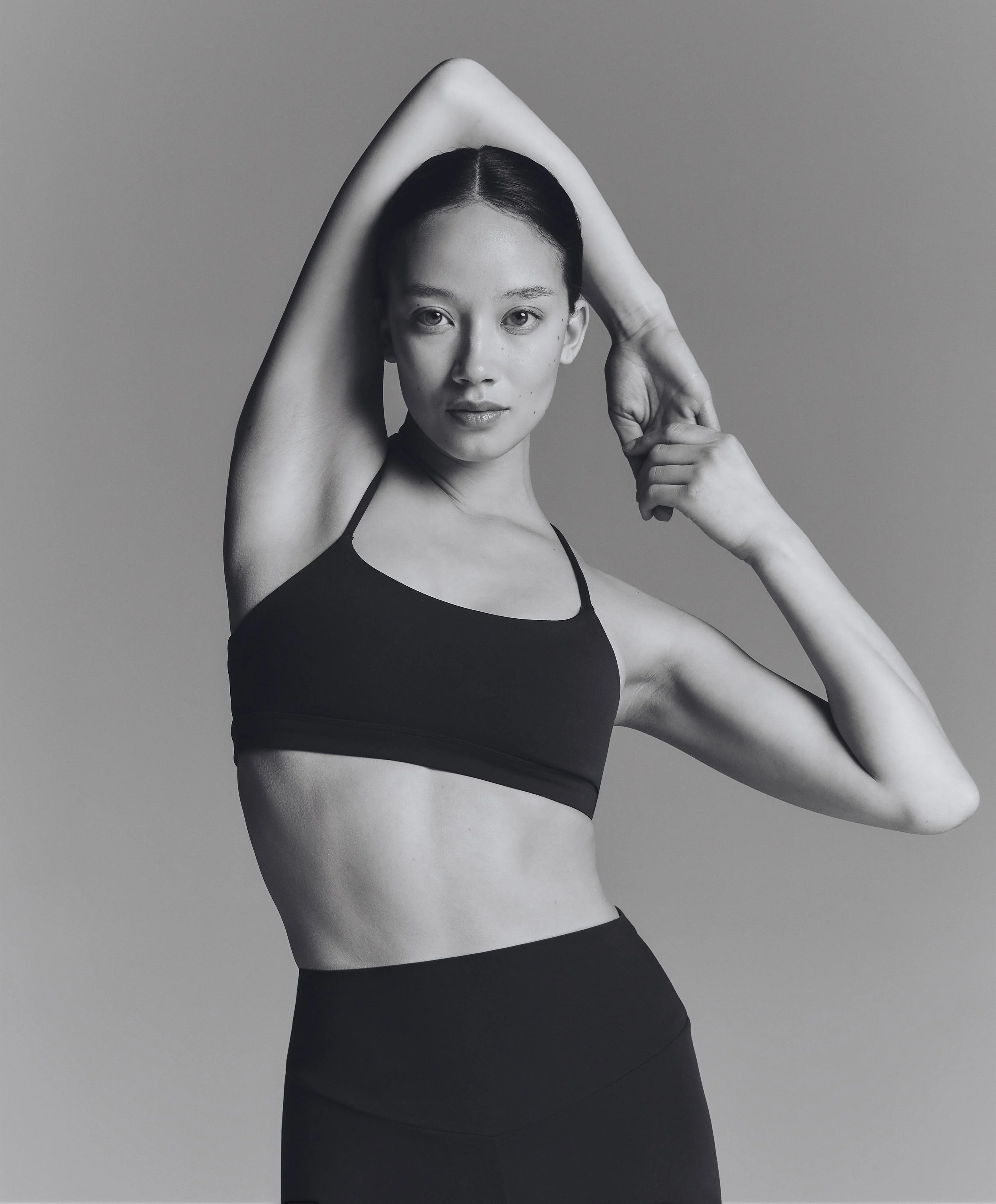 Black and white photo of a woman with short dark hair, wearing a sports bra and leggings, posing with her arms raised, one hand on her head, and the other touching her face, against a plain background.