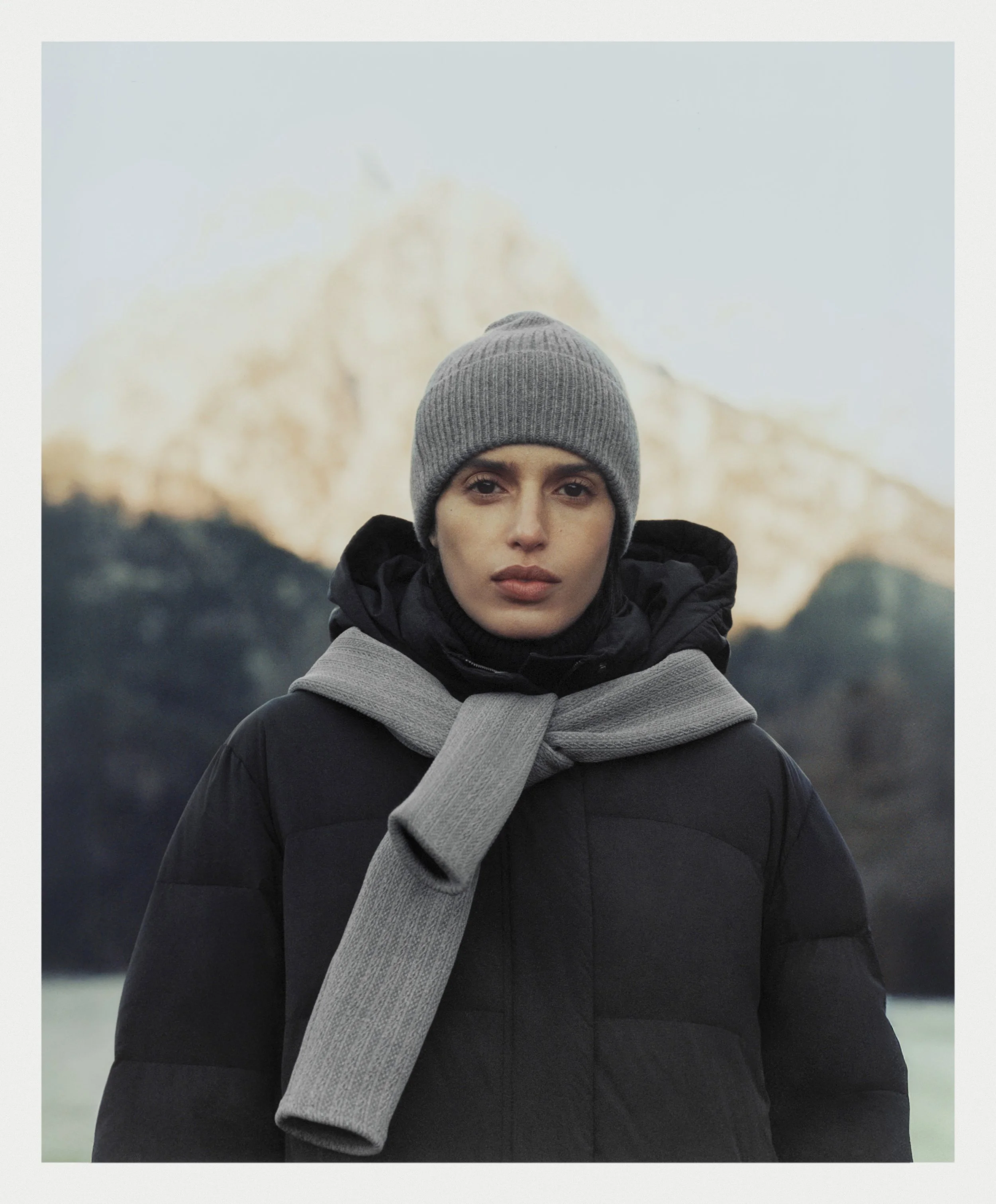 A woman wearing a gray beanie, black jacket, and gray scarf, standing outdoors with a mountain in the background
