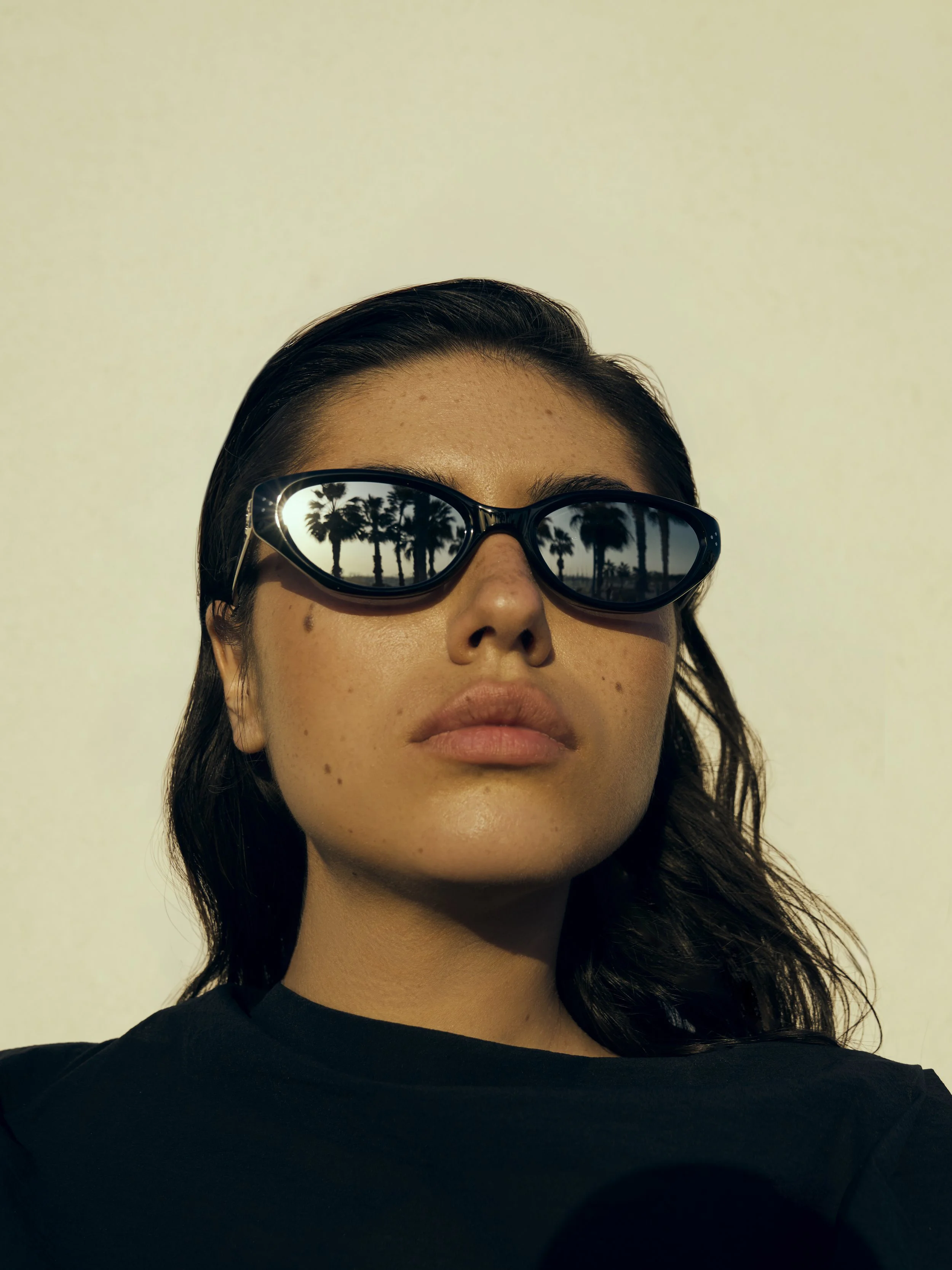A woman with dark hair wearing sunglasses that reflect palm trees and a sunset or sunrise sky, against a plain light-colored background.