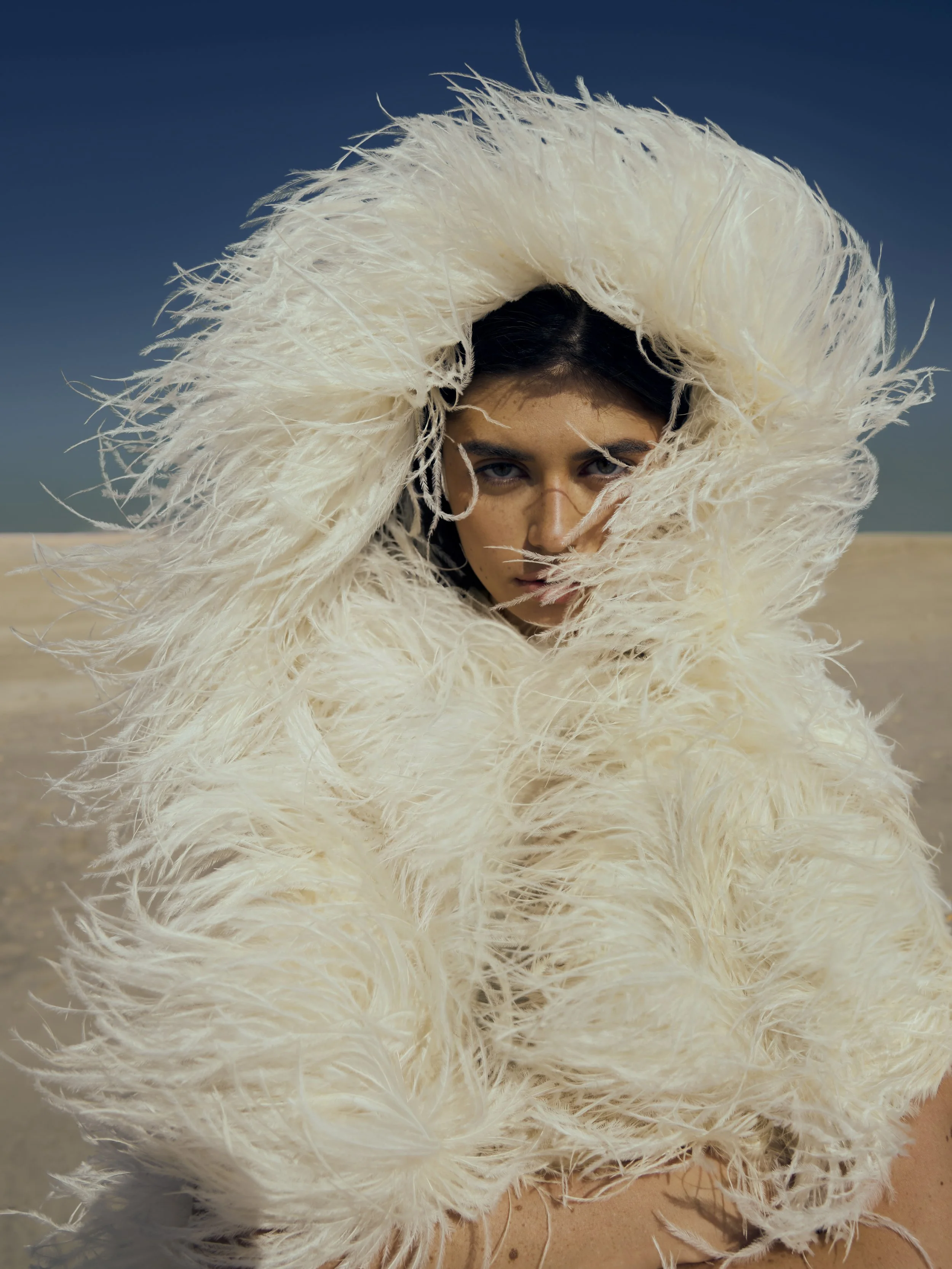 Person on a sandy beach wearing a large, fluffy white feathered hood or costume, with dark hair and partially visible face, under clear blue sky.