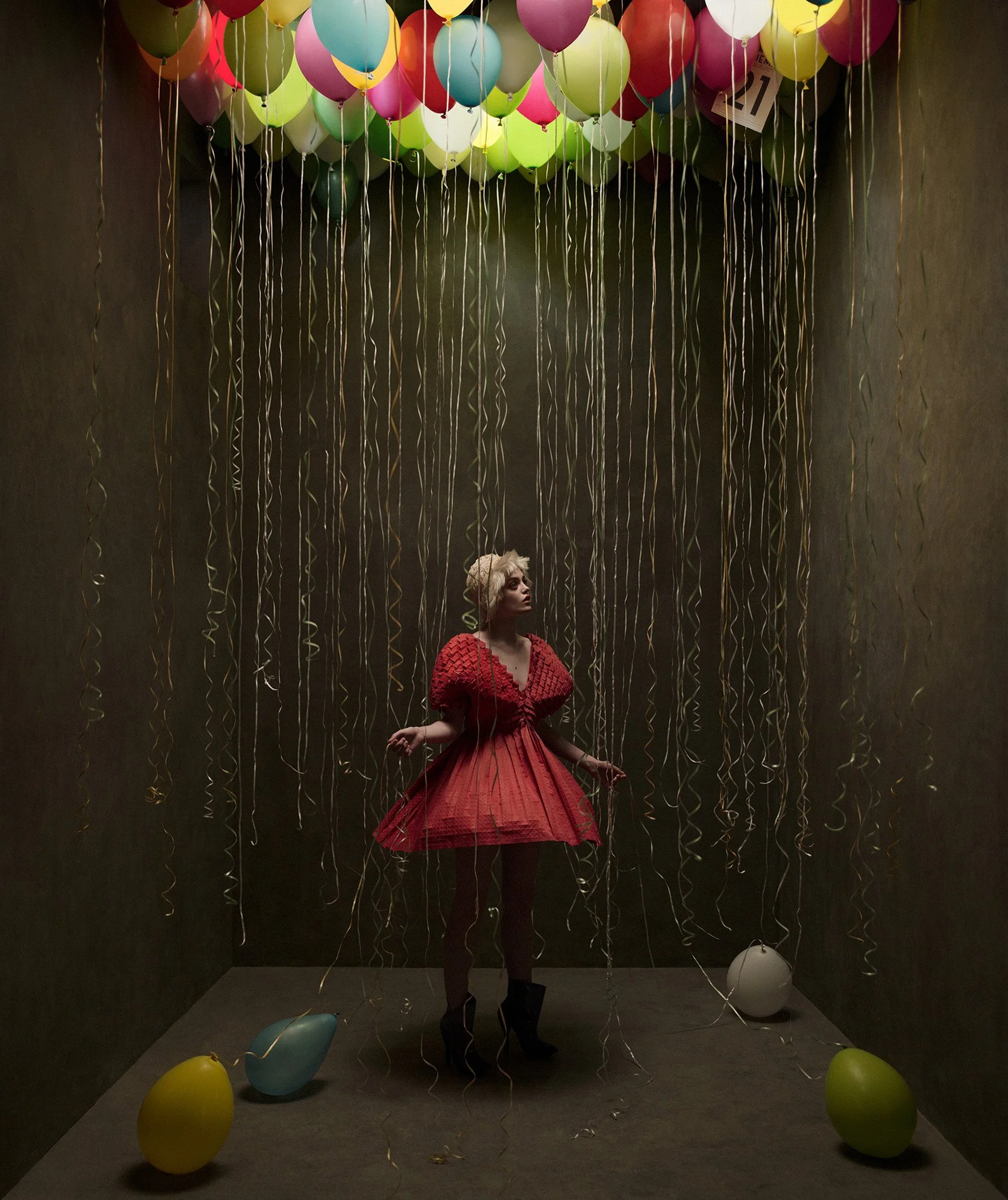 A woman in a red dress and black high heels is standing on a dark room floor surrounded by colorful balloons and streamers hanging from the ceiling. Some balloons are on the floor and others are floating above.