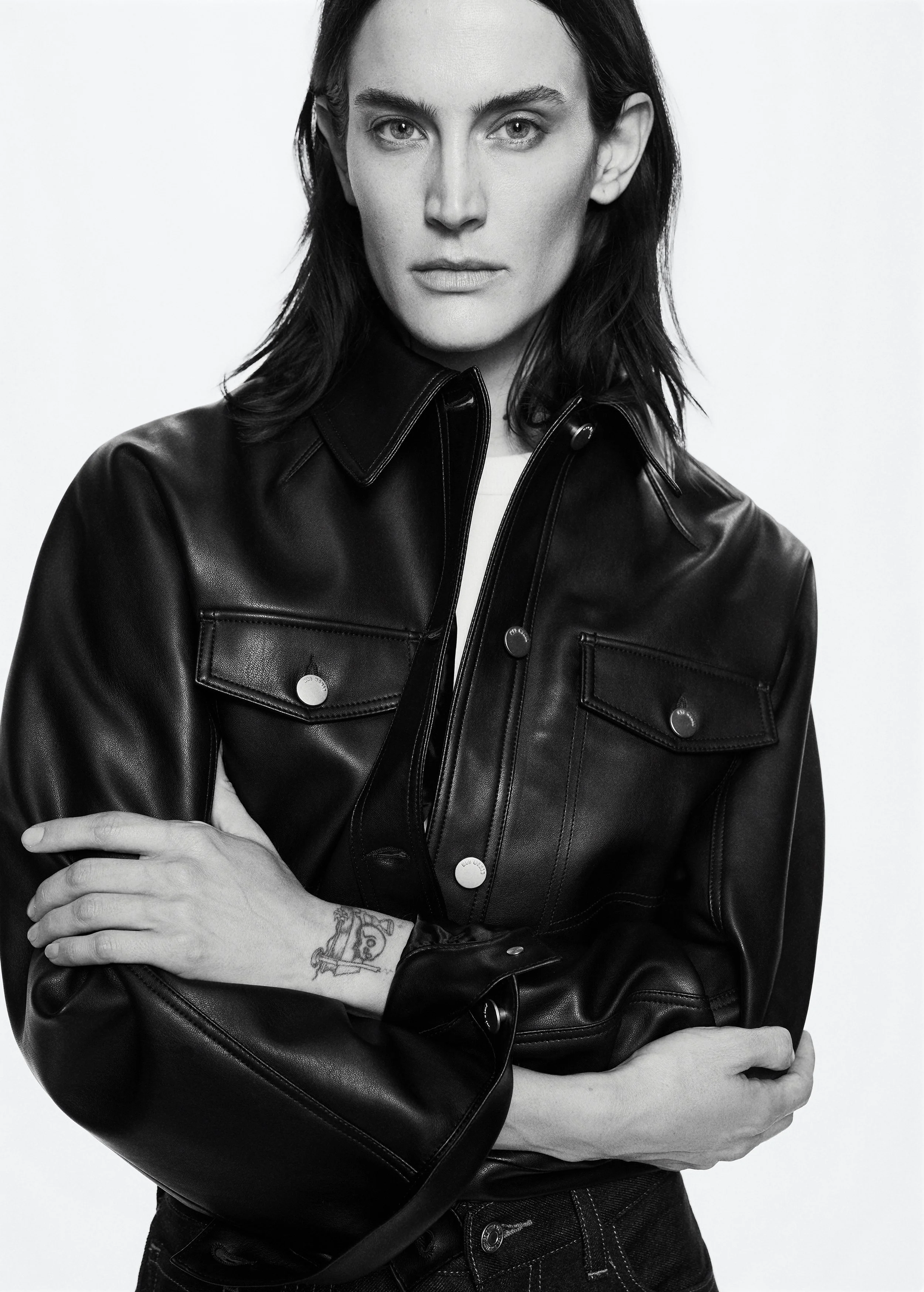 Black and white photo of a woman with dark hair wearing a leather jacket, looking directly at the camera with her arms crossed, showing a tattoo on her wrist.