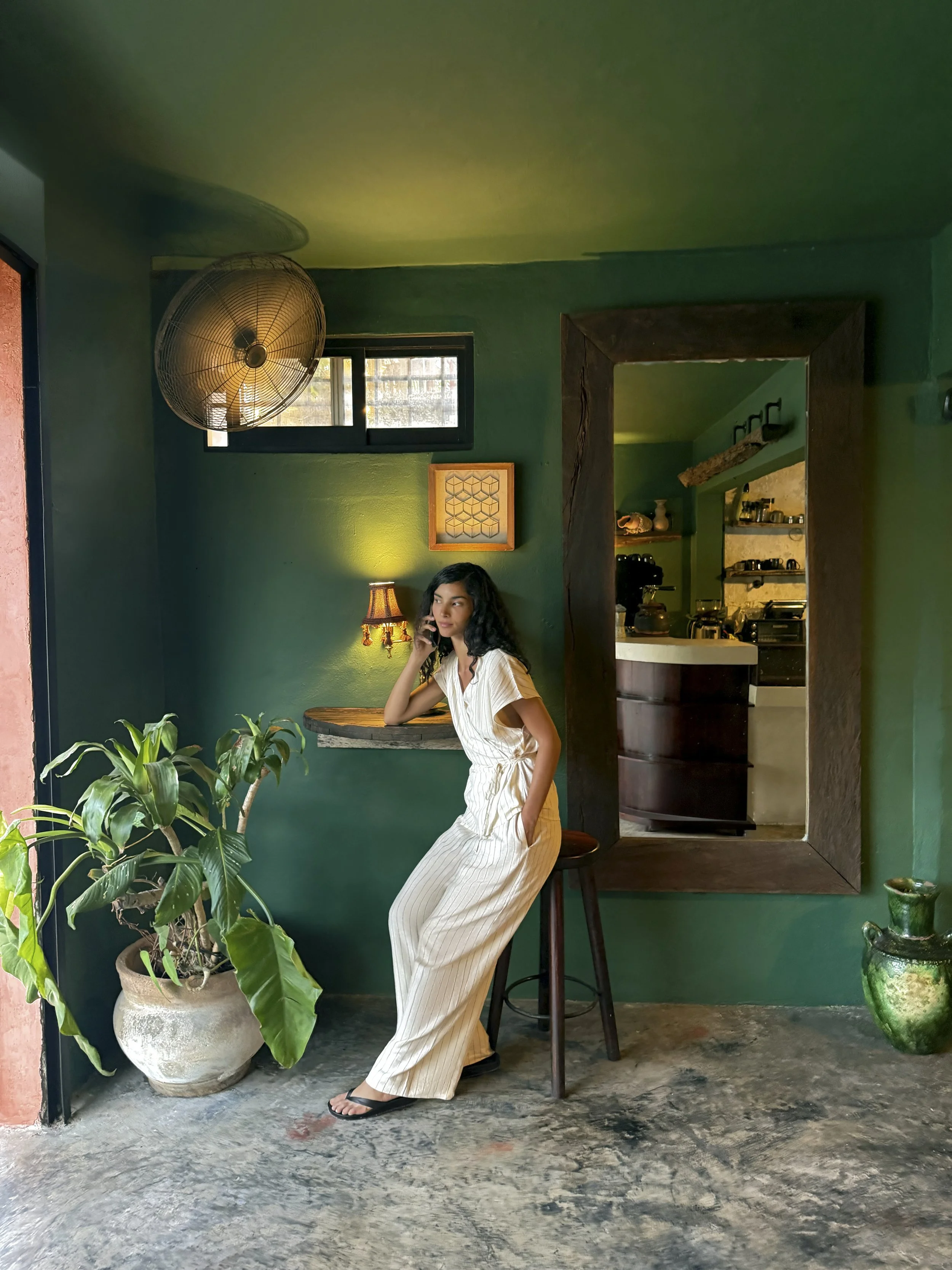 A woman with dark, wavy hair sitting on a bar stool in a cozy, green-walled cafe, talking on her mobile phone. She is wearing a light-colored, striped jumpsuit and black sandals. There is a large potted plant in the foreground, and a window, a wall l