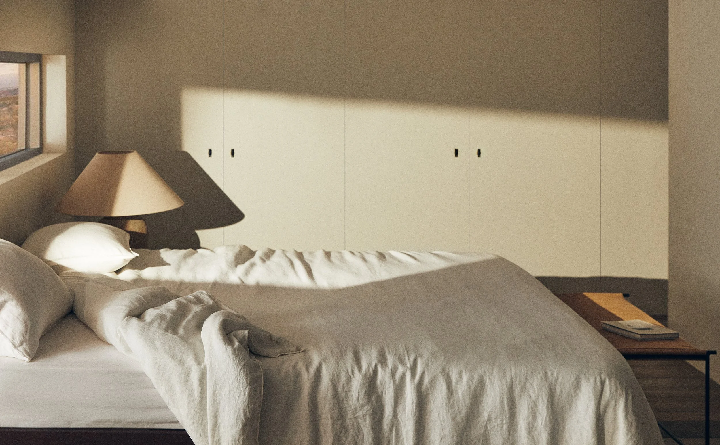A cozy bedroom featuring a bed with white linens and pillows, a soft beige lampshade on a side table, and a large built-in closet with closed white cabinet doors. There is natural sunlight casting shadows on the wall, and a small window showing a lan