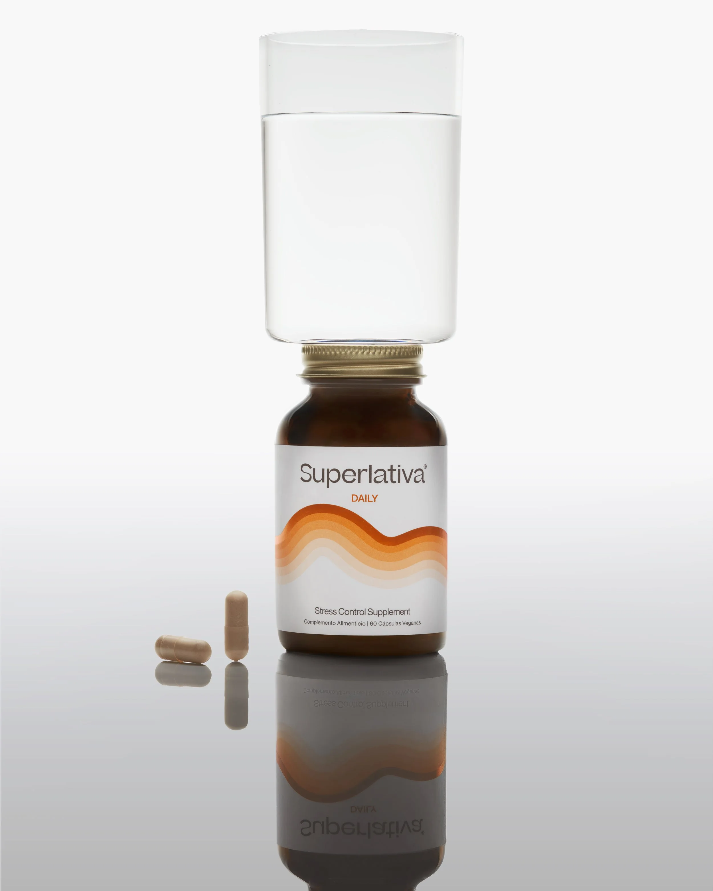 A brown bottle labeled 'Superlativa DAILY' with orange wave design, containing stress control supplement capsules, reflected on a glossy surface with two capsules beside it.