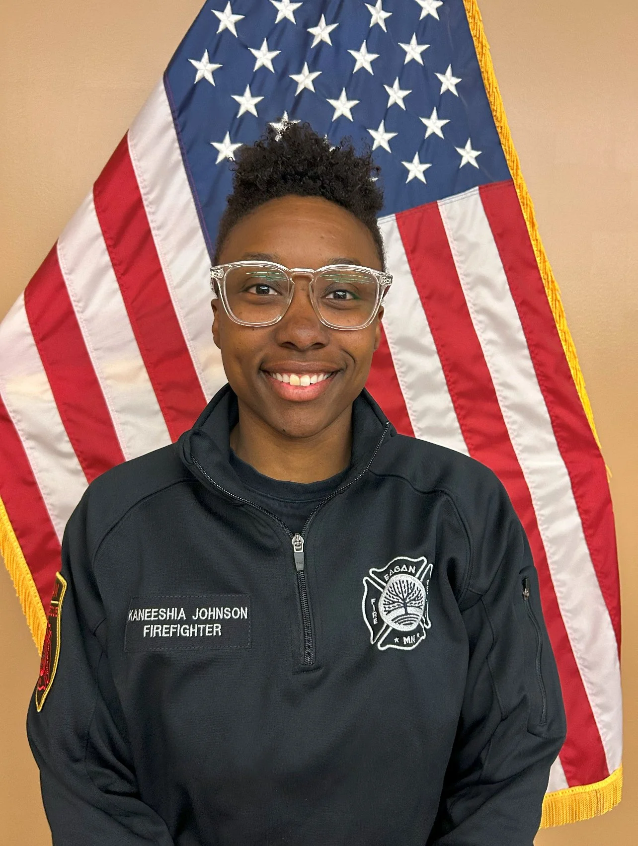 A smiling young African American woman in firefighter uniform standing in front of an American flag.