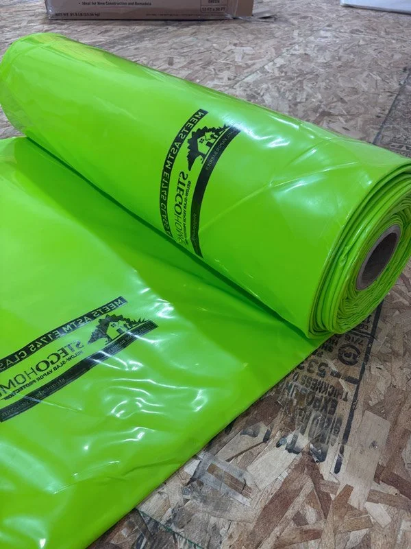 A bright yellow-green roll of plastic sheeting with black and yellow branding, placed on a wooden surface. The sheeting is partially unrolled, and the logo and text indicate it is from a concrete and masonry supplier.