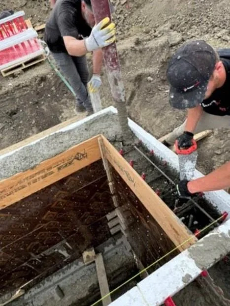 Two construction workers working on a foundation, one pouring concrete and the other using a tool, with wood framing around the opening.