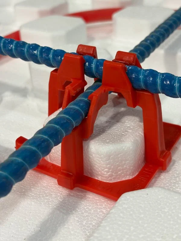 Close-up of metal rebar held in red plastic rebar chairs on a dirt surface, used in construction or concrete reinforcement.