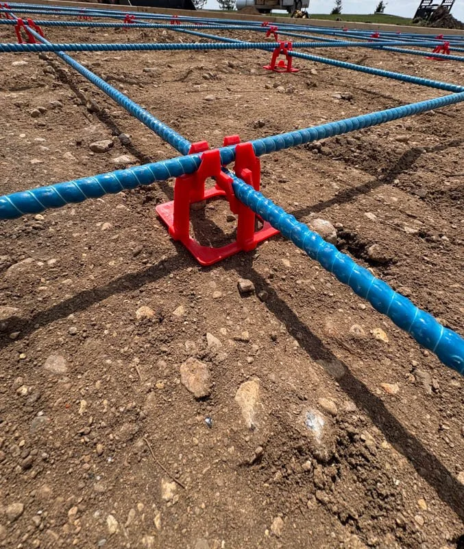 Construction site with blue and red rebar chairs arranged in a grid pattern on dirt ground.