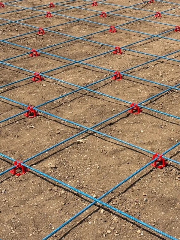 Metal rebar grid laid over dirt for concrete pouring, with red spacers at intersections.
