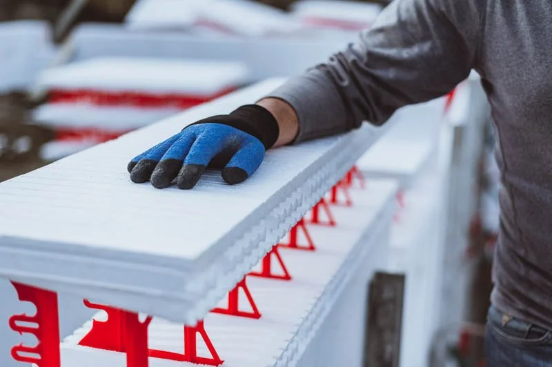 A worker wearing gloves and a gray long-sleeve shirt handling a white foam board at a construction site.