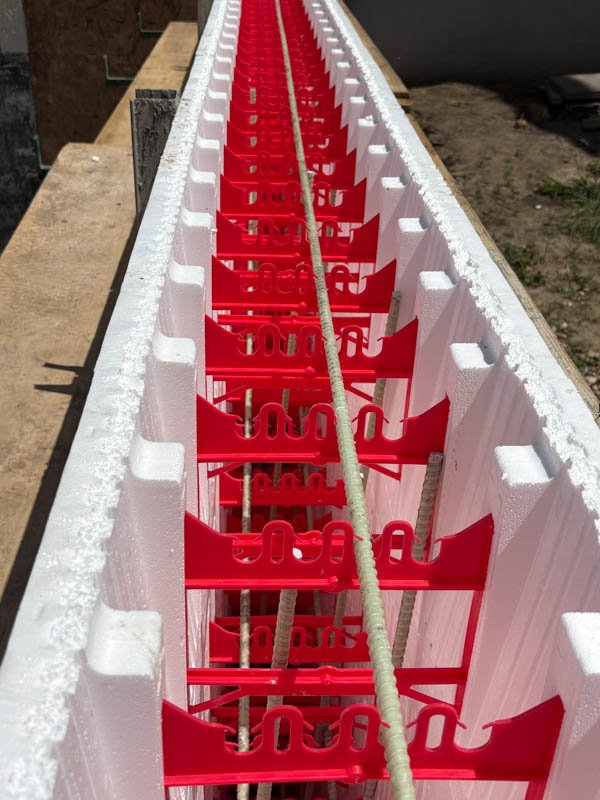 Long white styrofoam form with red plastic support beams and metal rebar inside, lying on a workbench at a construction site.