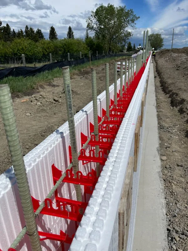 Construction site showing a concrete formwork with red spacers and steel reinforcing bars, part of a sidewalk or foundation under construction.