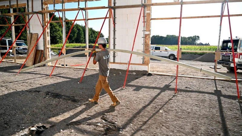 A construction worker wearing a helmet walks inside a partially built structure with red support beams, carrying a metal beam. Outdoors with a truck and open field visible in the background.