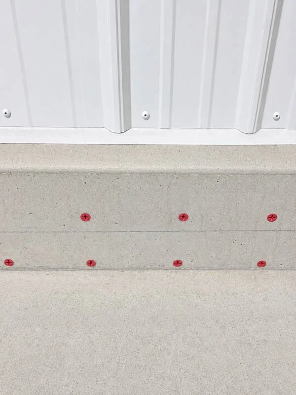 Close-up of a concrete floor with red anchors or screws arranged in a pattern near a white metal wall or panel.