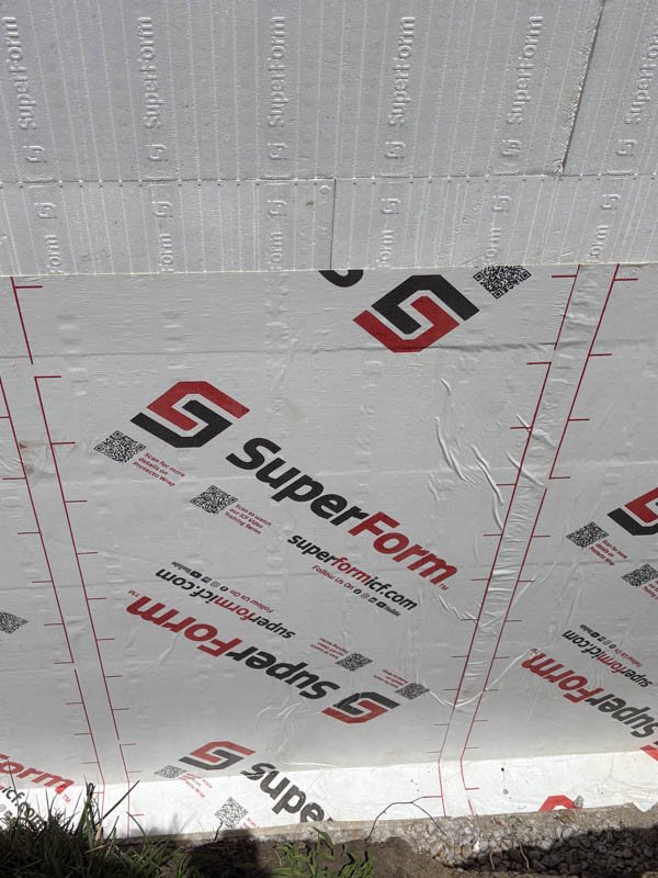The image shows a close-up view of a building's wall under construction, covered with white SuperForm insulation or house wrap material with the SuperForm logo and website printed on it.