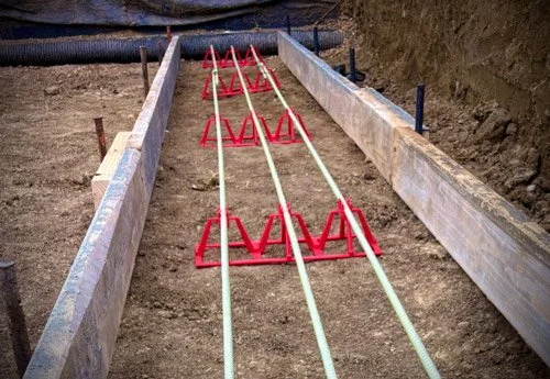 Construction site with wooden formwork and red runners for concrete pouring, with three white pipes in the center.