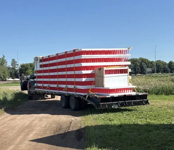 Flatbed trailer loaded with red and white striped barriers on a grassy area near a dirt road, with a truck attached and trees in the background.