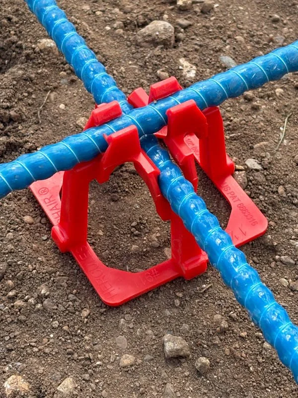 Close-up of metal rebar held in red plastic rebar chairs on a dirt surface, used in construction or concrete reinforcement.