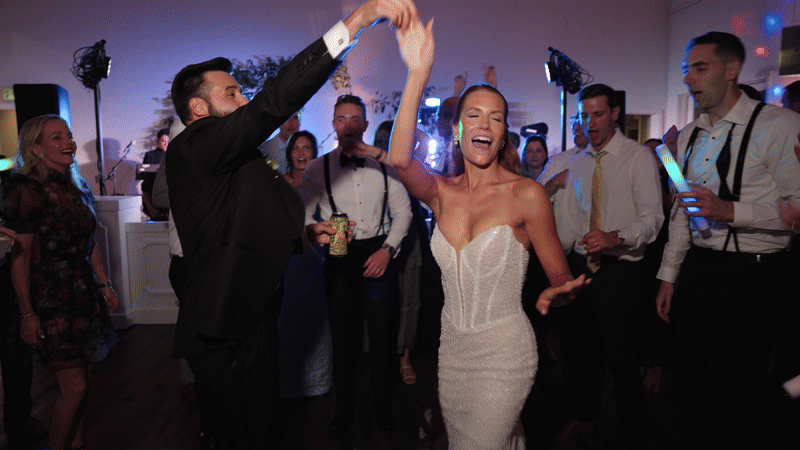 People dancing and celebrating at a wedding reception with a bride in a white wedding dress and groom in a tuxedo, surrounded by guests.