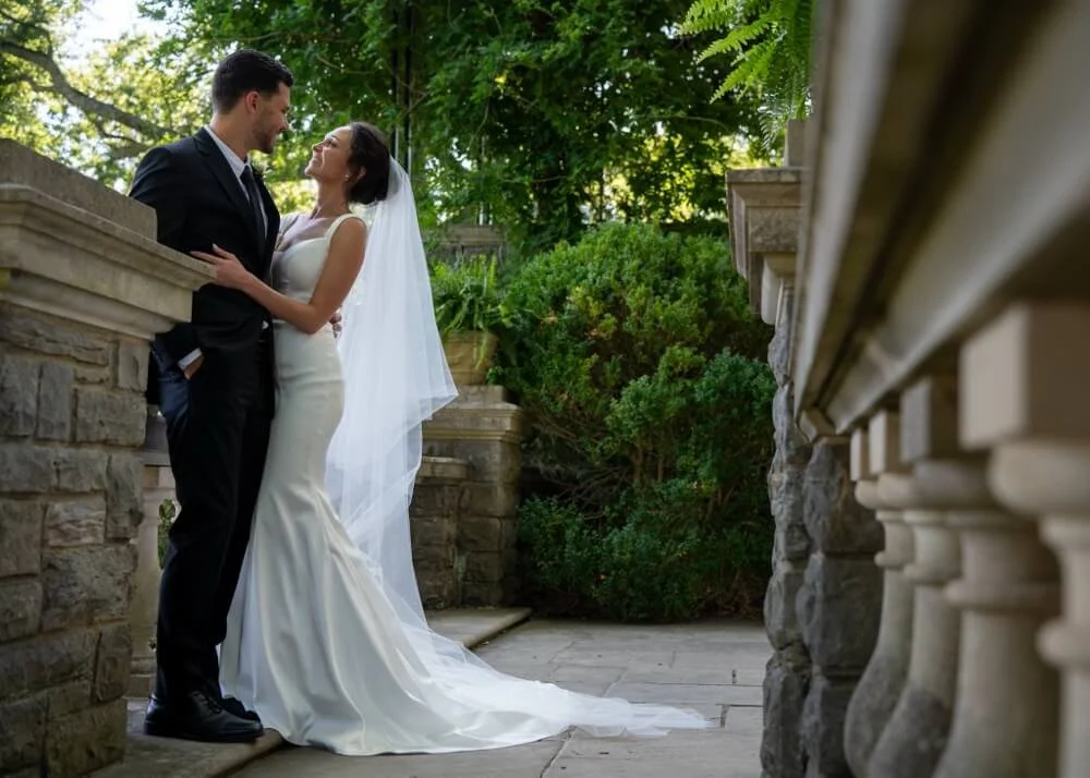 A bride and groom standing close together outdoors, gazing into each other's eyes, with lush greenery and stone architectural features in the background.