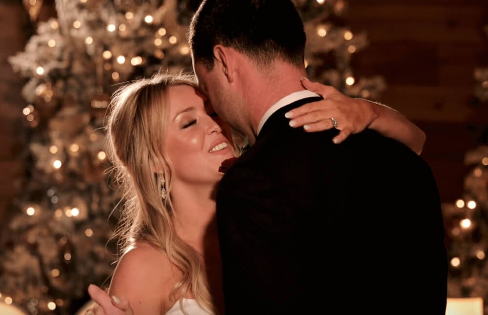 A smiling woman with long blond hair and a man with dark hair wearing a tuxedo share a close embrace in front of a decorated Christmas tree.