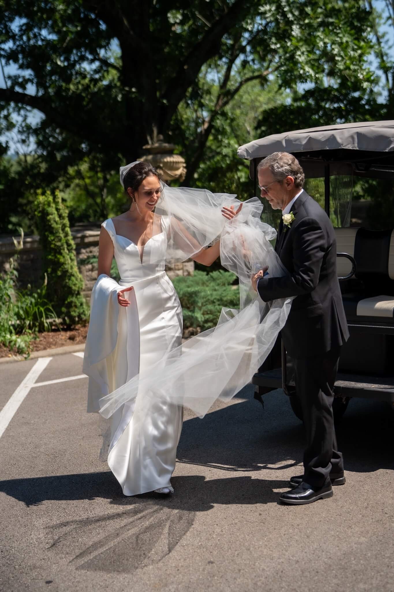 A bride in a white wedding dress and veil is smiling and laughing as an older man, possibly her father, dressed in a black tuxedo, helps her with her wedding veil outside in a sunny garden setting. There is a golf cart nearby and large trees in the background.