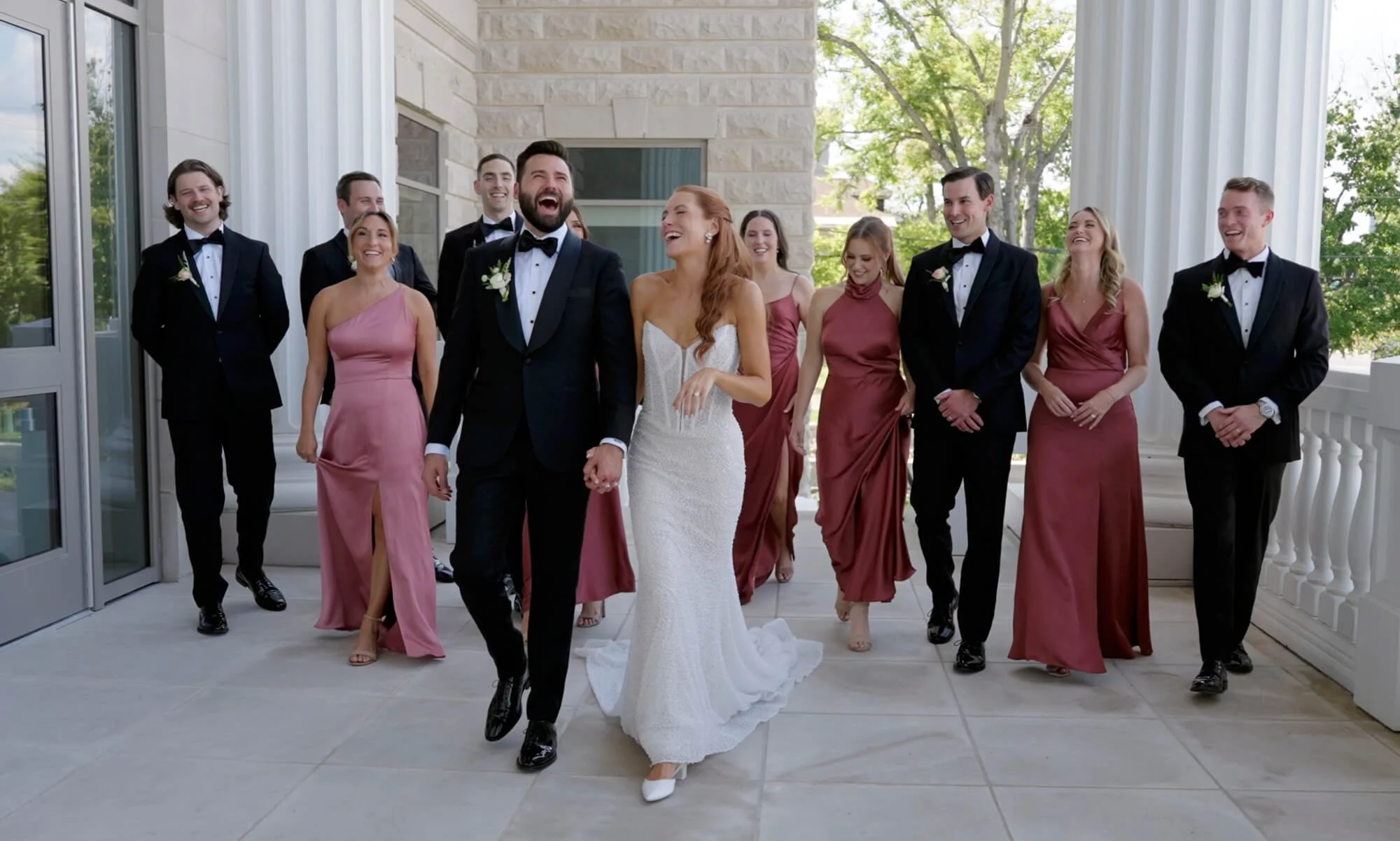 A bride and groom walking hand-in-hand with their wedding party, smiling and laughing, on a porch with white columns and green trees outside.