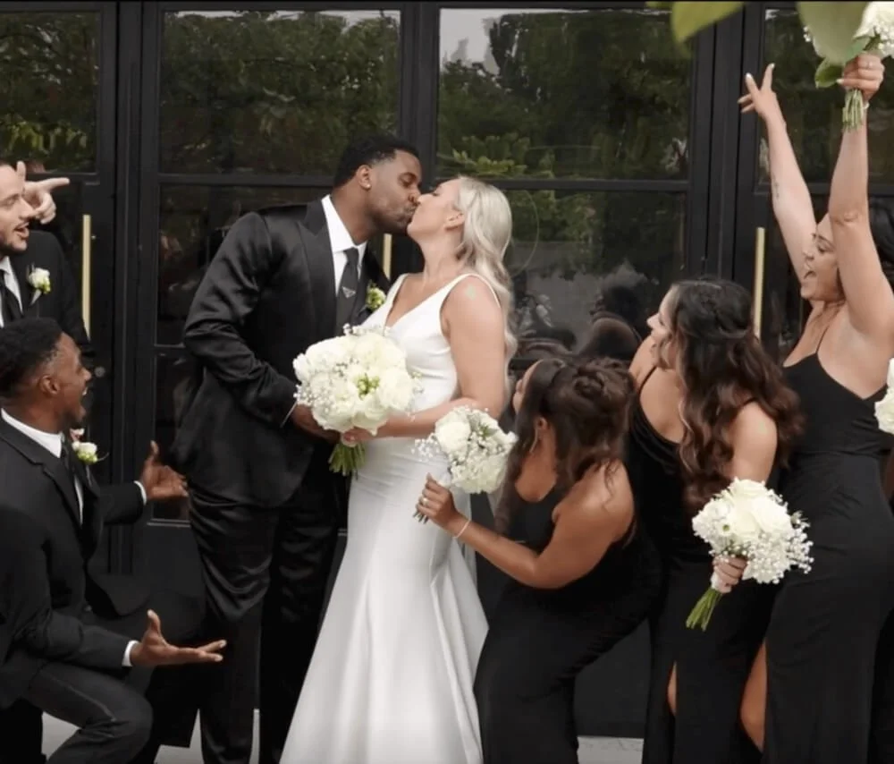 A wedding celebration with a bride and groom kissing, surrounded by wedding party members, some holding bouquets and celebrating.