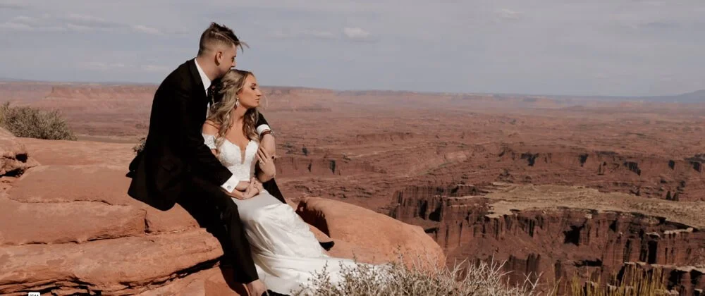 A couple dressed in wedding attire sitting on a rock ledge in the Grand Canyon, overlooking the canyon landscape.