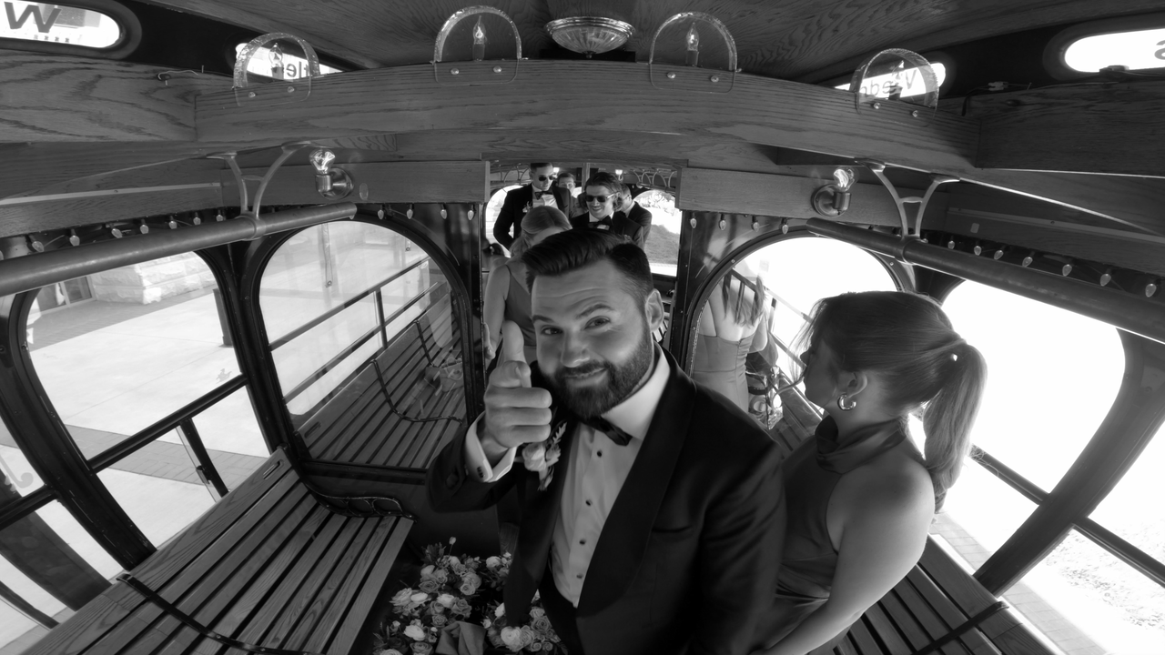 Black and white photo of a man in a tuxedo giving a thumbs-up inside a vintage trolley with several people behind him.