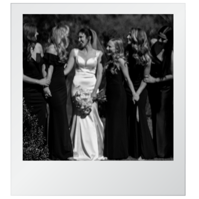 Black and white photo of a bride in a wedding gown holding a bouquet, surrounded by her bridesmaids in matching dresses, all standing outdoors and holding hands.