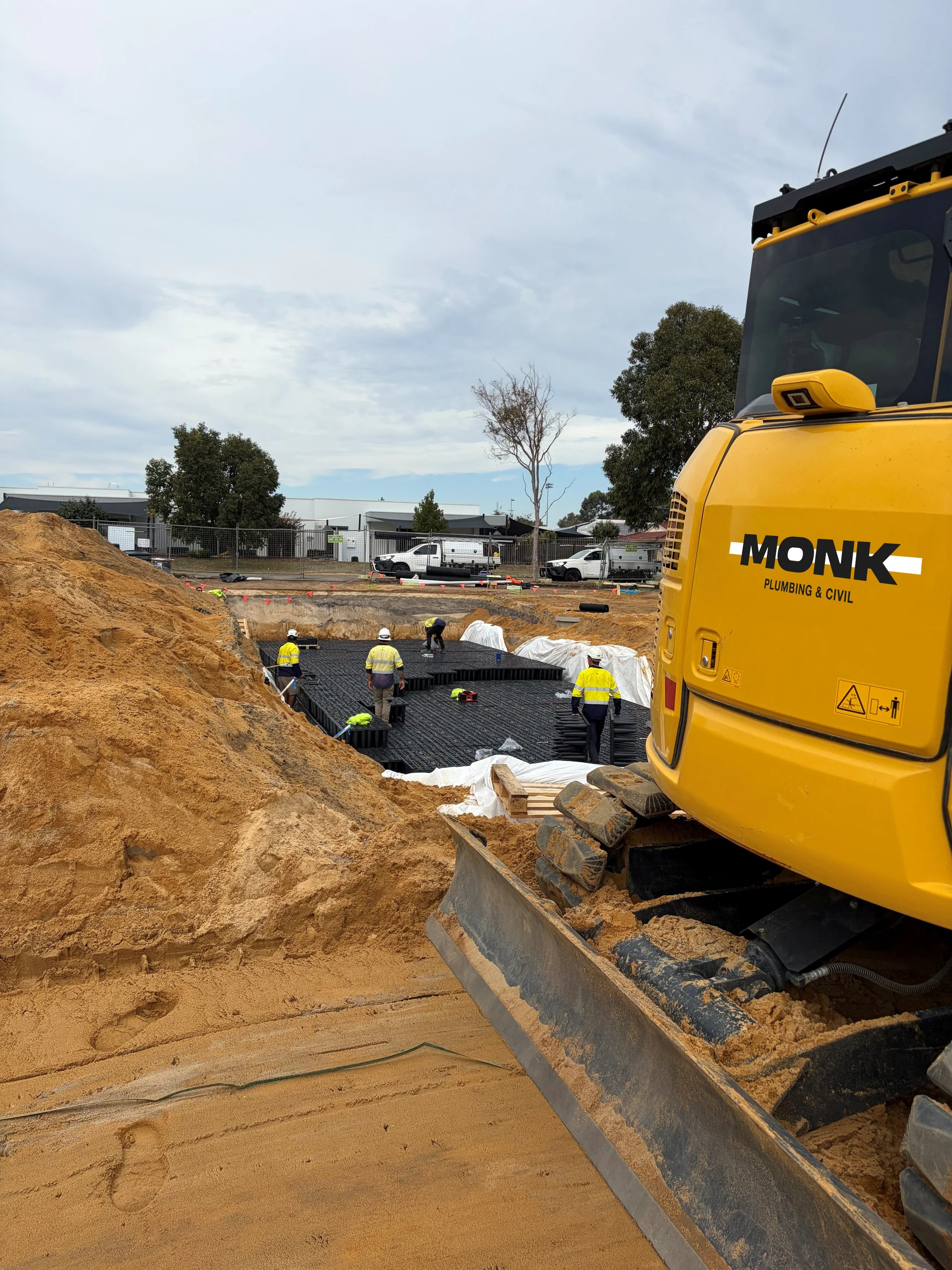 Construction site with workers installing a foundation, a yellow excavator labeled 'MONK Plumbing & Civil,' and a partially covered area in the background with vehicles parked nearby.