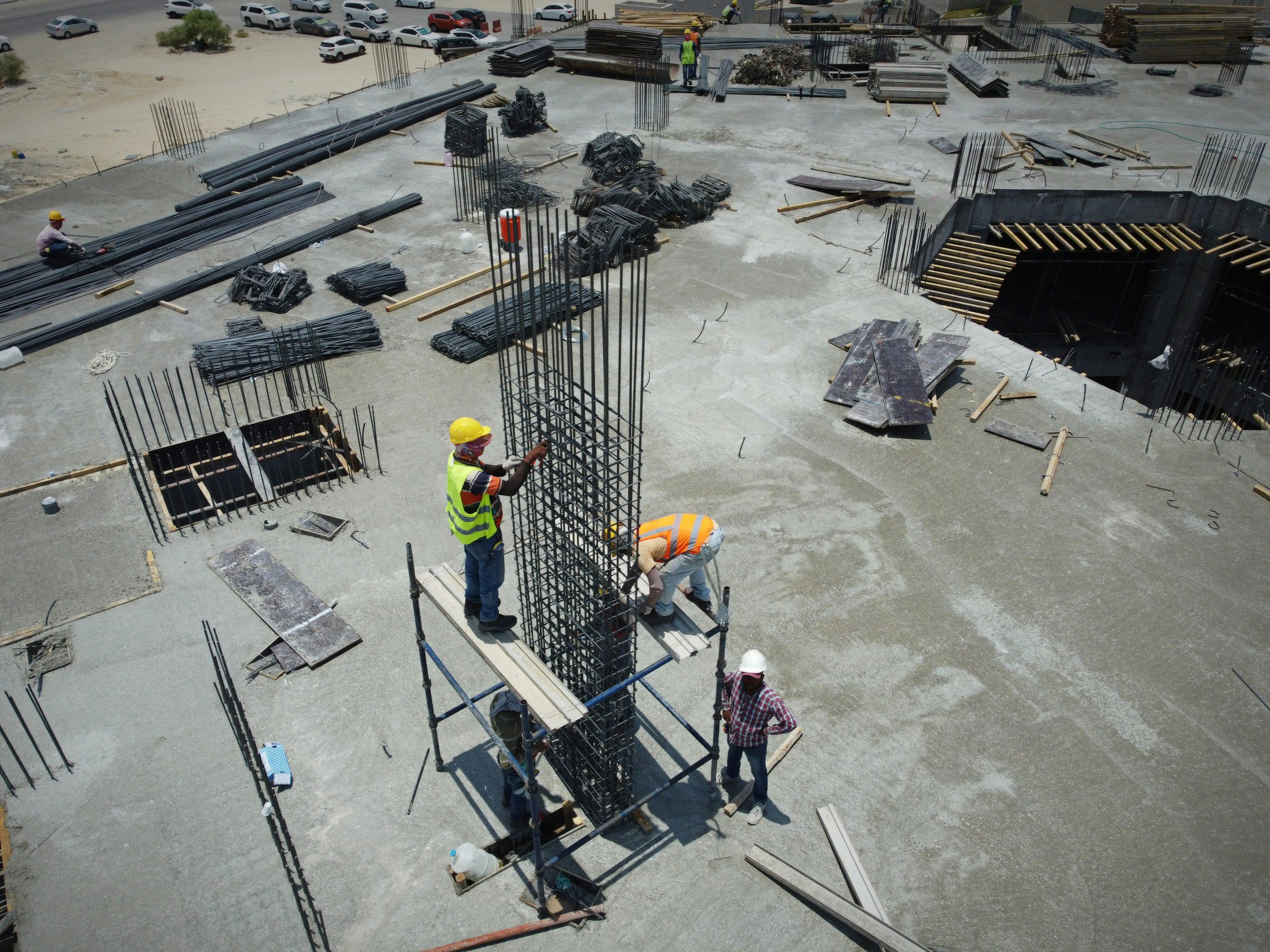 Construction workers on a building site, incorporating steel rebar into the concrete foundation, with construction materials and vehicles in the background.