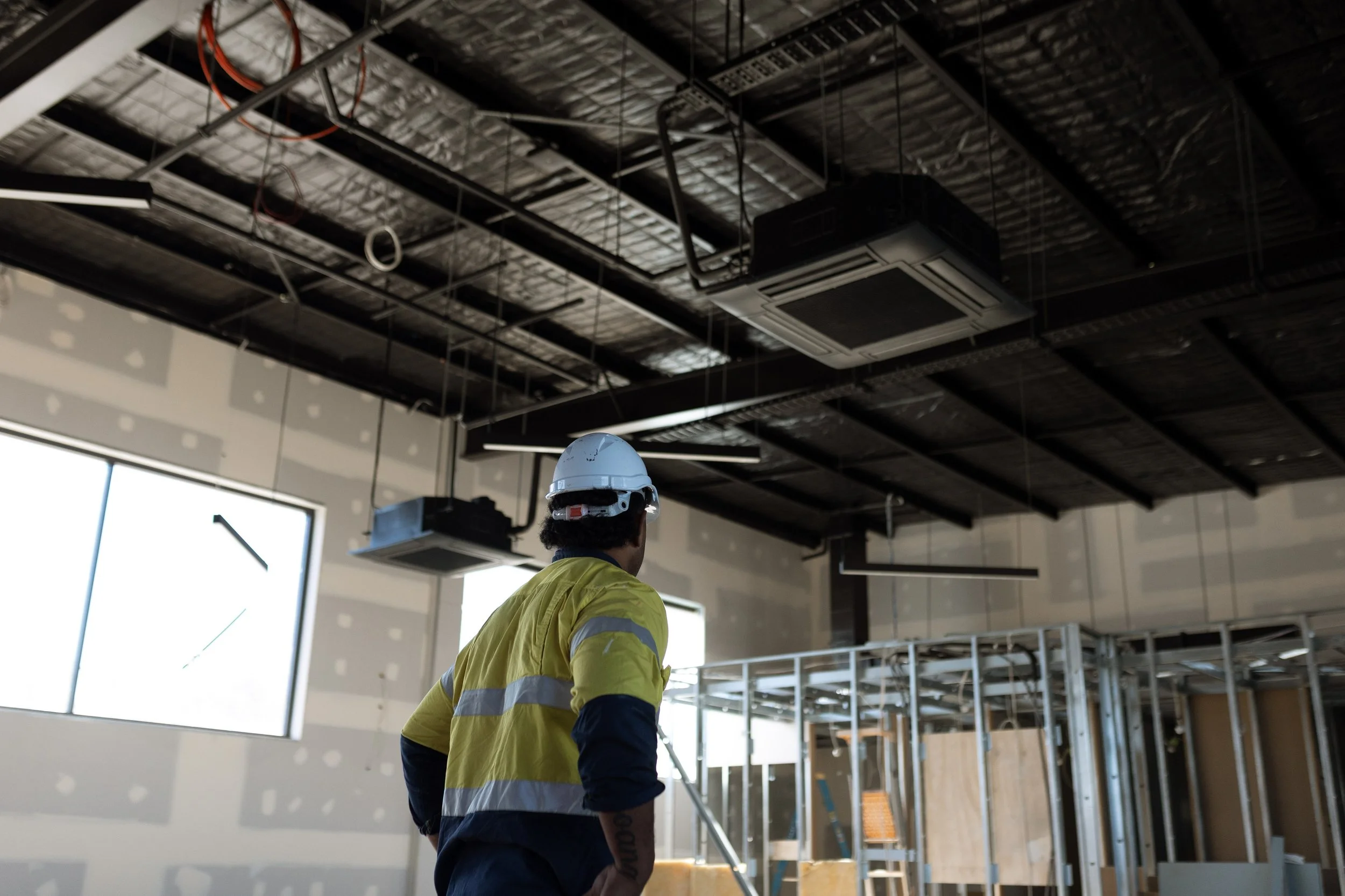 Construction worker wearing a yellow safety vest and white hard hat inside a building under construction with exposed ceiling and metal framework.