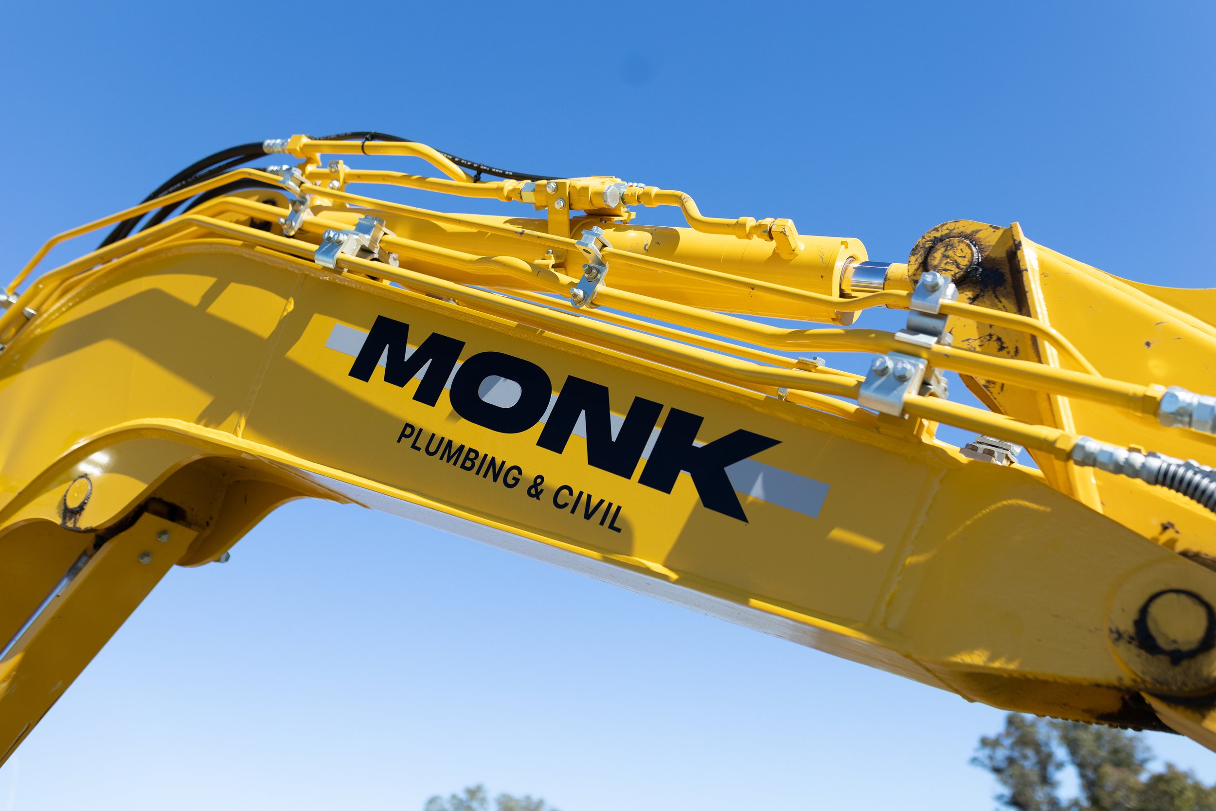 Close-up of a yellow excavator arm with hydraulic hoses and metal joints against a clear blue sky, with part of a company logo visible on the arm.