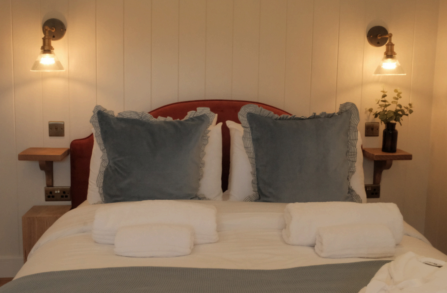 A neatly made bed with two large gray pillows with ruffled edges, two white pillows, and rolled white towels on top of a beige bedspread. There are two wall-mounted lamps with exposed bulbs on either side, a small wooden bedside table on each side, and a dark vase with a plant on the right side.